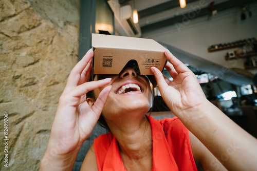 Fototapeta Naklejka Na Ścianę i Meble -  A young Indian Asian woman sitting in a cafe tries VR (Virtual Reality) goggles for the first time and is surprised and delighted. She is smiling and looking up.