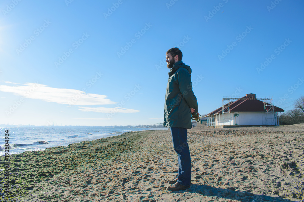 a handsome pensive young man with a beard stands on the seashore and looks into the distance. dreaming guy on the background of the sea. sunny day