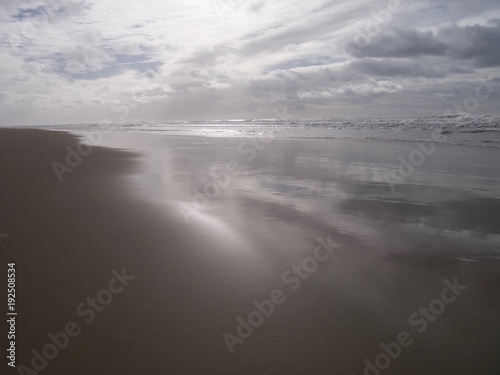 Stormy sky, at sunset, reflected on the surface of the water tide on a sandy Atlantic beach with the sun illuminating white waves, in winter in South West France, near Lacanau