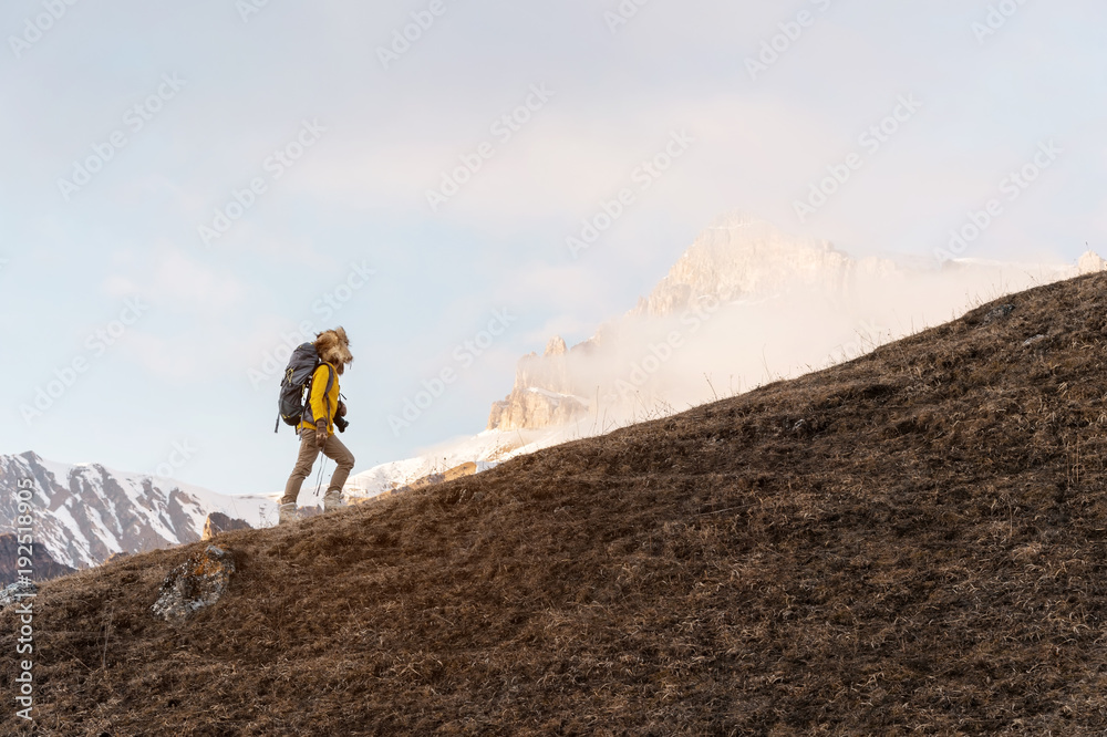 A backpacker in a big fur hat and gloves with a backpack on his back goes uphill against the background of epic cliffs in the clouds. The concept of mountain tourism in any season