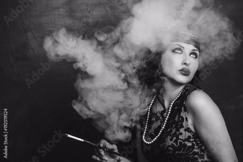 brunette girl in black dress in the style of decadence holds the mouthpiece and blows smoke © Sonya