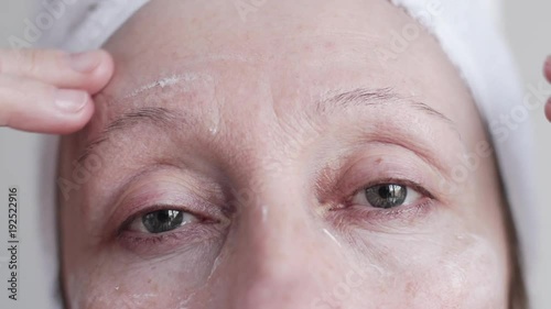 Slow motion shot of beautiful middle-aged caucasian woman in bathroom look at camera and applying cosmetic cream on face. photo