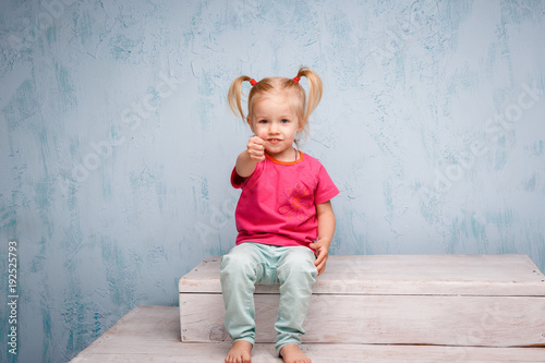 Little funny blue-eyed girl child blonde with a haircut two ponytails on her head sitting on a gossip on the background of an old textured wall in blue. Dressed in stylish bright clothes