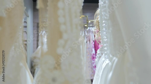 Beautiful young bride standing in a bridal boutique. Smiling woman shopping for wedding outfit in bridal clothing store. Young woman looking at bridal dresses. Pretty woman is preparing for wedding.