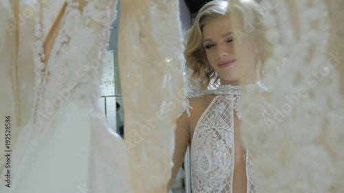 Beautiful young bride standing in a bridal boutique. Smiling woman shopping for wedding outfit in bridal clothing store. Young woman looking at bridal dresses. Pretty woman is preparing for wedding.