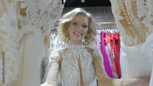 Beautiful young bride standing in a bridal boutique. Smiling woman shopping for wedding outfit in bridal clothing store. Young woman looking at bridal dresses. Pretty woman is preparing for wedding.