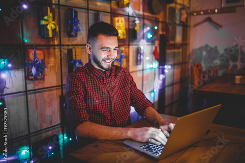Theme training and computers. A young man with a beard and in a shirt uses a laptop, prints on the keyboard in a coffee shop at a wooden table in the evening. Christmas decor and hang a garland © Elizaveta
