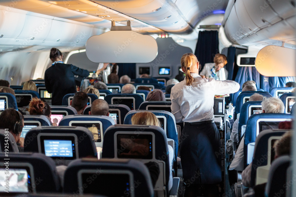 Fototapeta premium Wnętrze dużego samolotu komercyjnego z pomocnikami lotniczymi obsługującymi pasażerów podczas lotu. Stewardesy w ciemnoniebieskim mundurze idącym do ołtarza. Skład poziomy.
