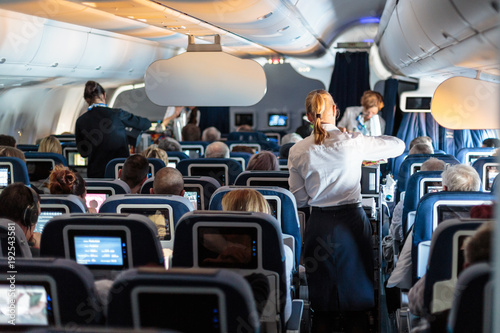 Interior of large commercial airplane with flight attandants serving passengers on seats during flight. Stewardesses in dark blue uniform walking the aisle. Horizontal composition.