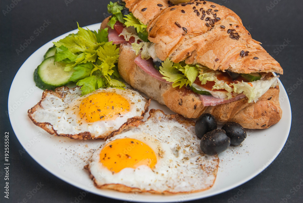 Breakfast with Fried Eggs and Sandwich Croissant on White Plate on black Background.