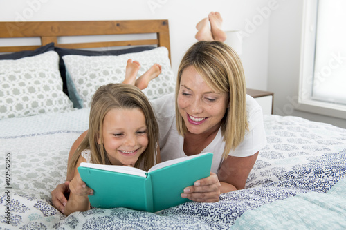 young blond Caucasian mother lying on bed with her young sweet  7 years old daughter reading book together at home bedroom
