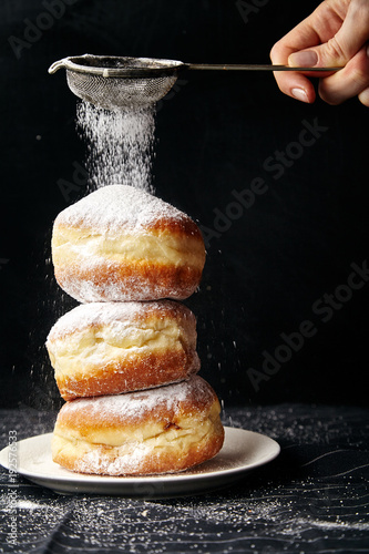 A stack of three sufganiyot donuts with jelly on black background. Female hand is covering them with sugar powder