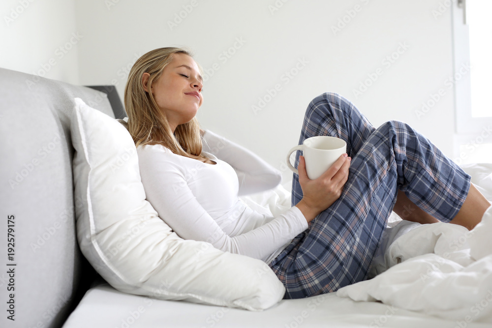 Hübsche blonde Frau im Schlafanzug und mit einer Tasse in der Hand in ihrem Bett lächelt Hübsche blonde Frau im Schlafanzug und mit einer Tasse in der Hand in ihrem Bett lächelt