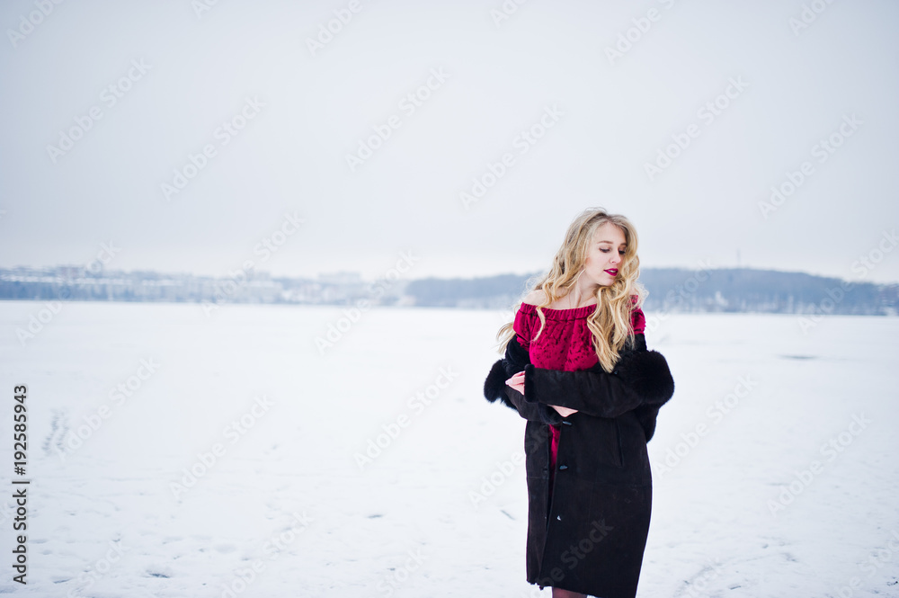 Elegance blonde girl in fur coat and red evening dress posed at winter snowy day.