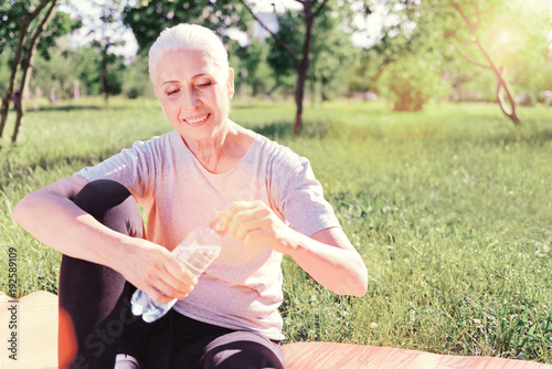 Resting outdoors. Positive elderly woman sitting on the ground pad while opening a bottle with water and expressing cheer