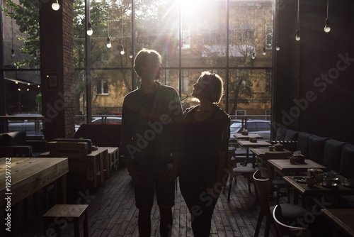 Nice couple standing on cozy empty cafe in loft style looking to each other with sunlight back view