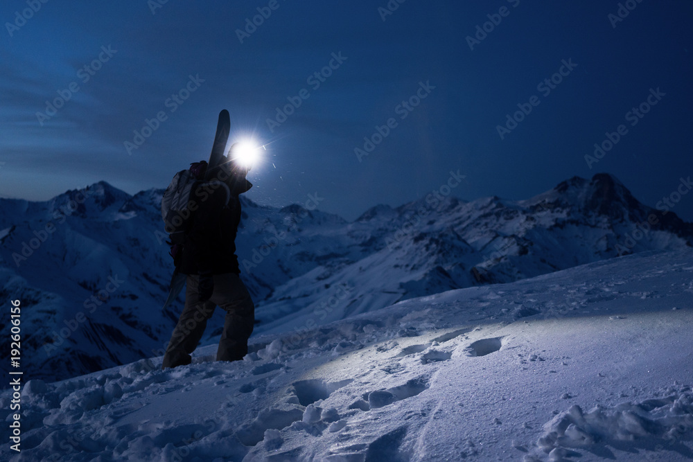 Professional tourist commit climb on great snowy mountain at night. Wearing backpack, headlamp and ski wear. Backcountry. Brave extreme traveler with a snowboard behind his back climbs winter slope - obrazy, fototapety, plakaty 