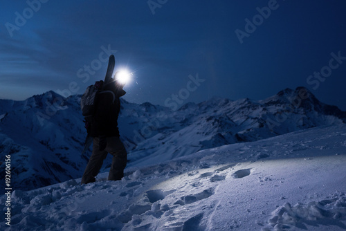 Professional tourist commit climb on great snowy mountain at night. Wearing backpack, headlamp and ski wear. Backcountry. Brave extreme traveler with a snowboard behind his back climbs winter slope