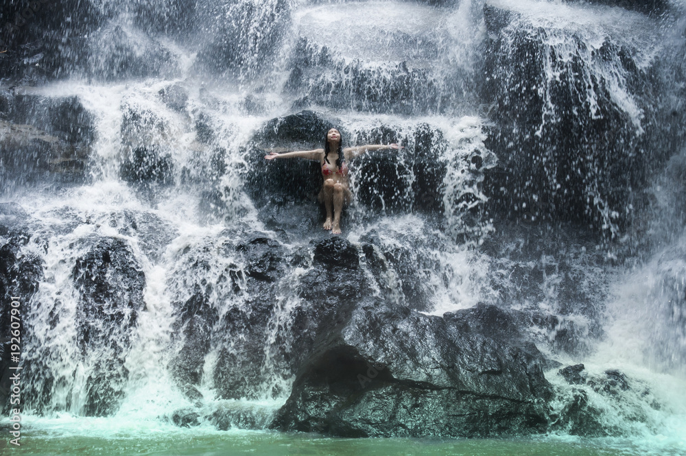 Obraz premium young beautiful and sweet Asian woman in bikini getting body wet under stream of natural amazing waterfall sitting on rock feeling purity