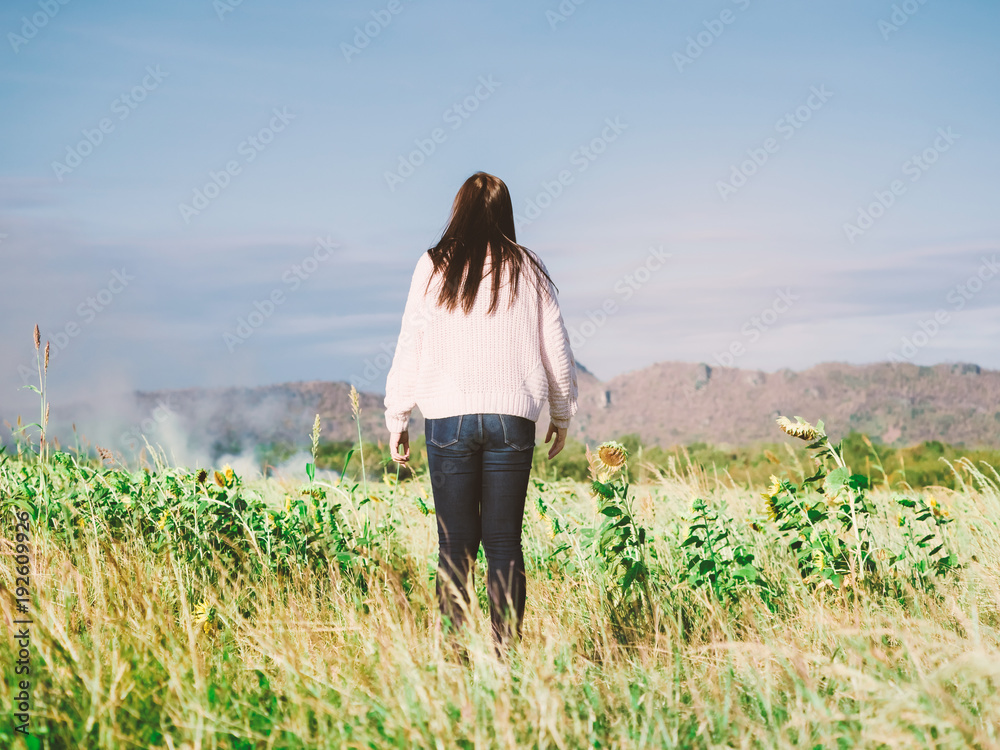 back side from young long hair woman with winter cloth stand and chill out at side of sunflower field on windy and cold weather from her holiday and vacation trip with soft focus background mountain