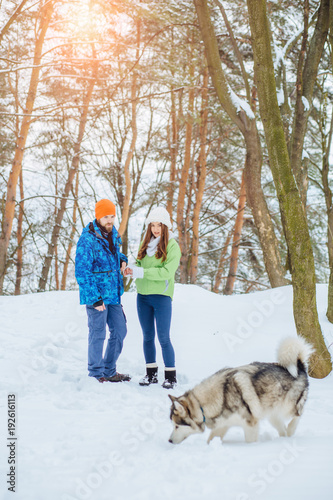 Sporty cute hipster couple in bright winter sportswear having fun in winter park with their friend husky alaskan malamute dog on a day talking with each other and smiling.