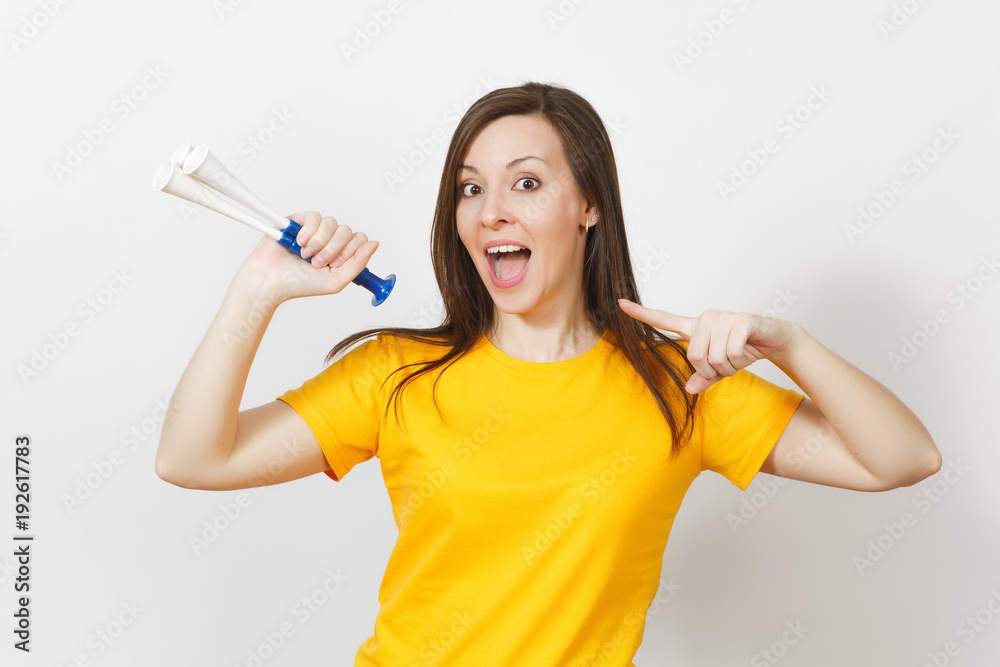 Beautiful European young happy brown-haired woman holding, blowing in football pipe, dressed in casual clothes, yellow t-shirt with shy charming smile isolated on white background. Sport fan concept.