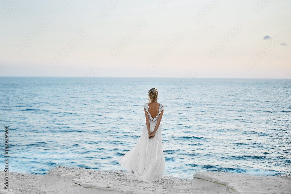 Beautiful young blonde model girl in white dress stands back and looks at the sea Beautiful young blonde model girl in white dress stands back and looks at the sea