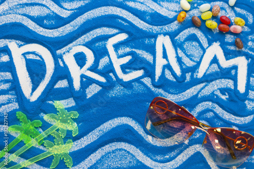 Dream inscription on a blue colored sand with waves, candies, sunglasses, top view, flat lay