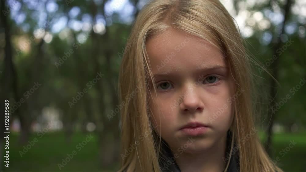 Serious little girl with light hair looking at camera while standing in a park on a cloudy summer day. Handheld slow motion close up shot Serious little girl with light hair looking at camera while standing in a park on a cloudy summer day. Handheld slow motion close up shot