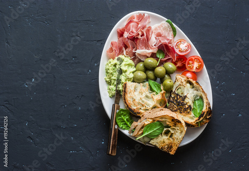 Snack bowl - prosciutto, olives, cherry tomatoes, avocado dip, grilled mozzarella spinach sandwiches on dark background, top view. Mediterranean style snack, appetizer, tapas. Free space