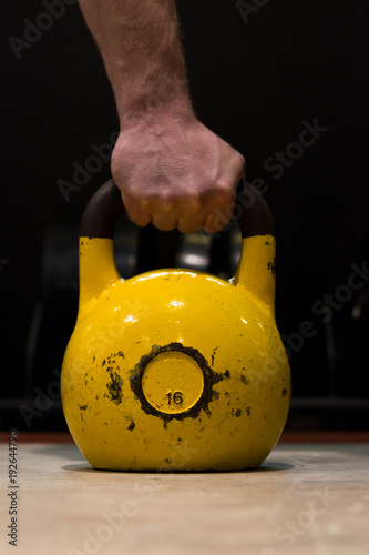 Strong human hand holding worn out yellow kettlebell in a gym on a wooden floor with black background