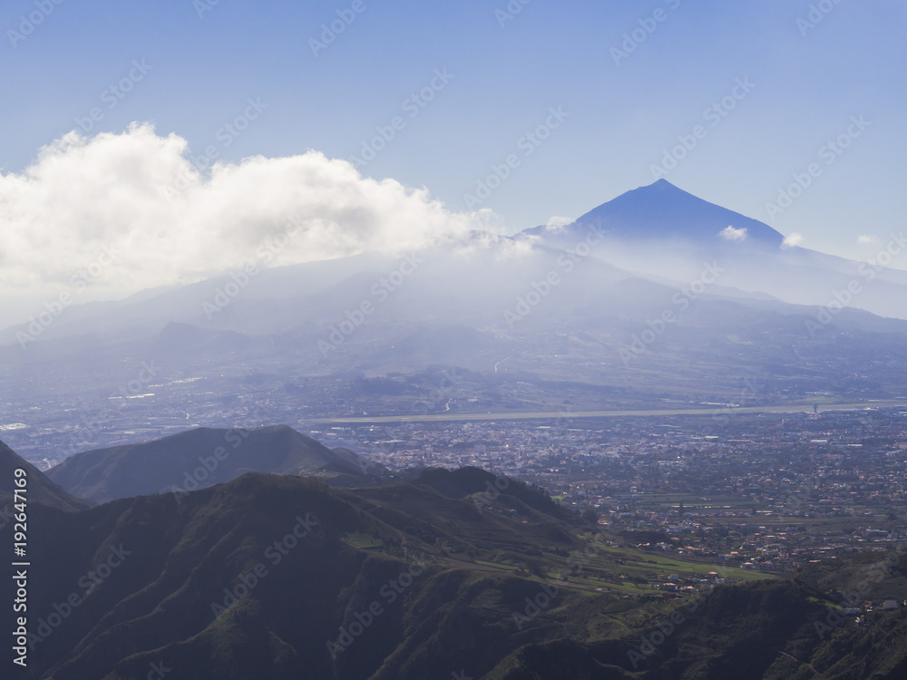 blue summit of volcano pico del teide highest spanish mountain in clouds with view on la orotava city and green hills, tenerife canary island, blue sky background