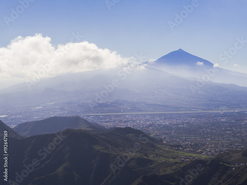 blue summit of volcano pico del teide highest spanish mountain in clouds with view on la orotava city and green hills, tenerife canary island, blue sky background