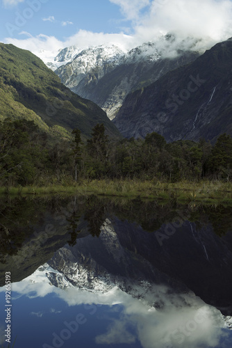 Paisaje de picos de montañas nevados y verdes con cielo nublado reflejado en un lago en Nueva Zelanda