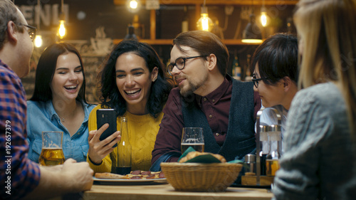 Beautiful Hispanic Woman Shows Interesting Stuff on Her Smartphone to Her Friends while They Have Good Time in Bar. They Laugh  Joke  Drink in Stylish Hipster Bar Establishment.