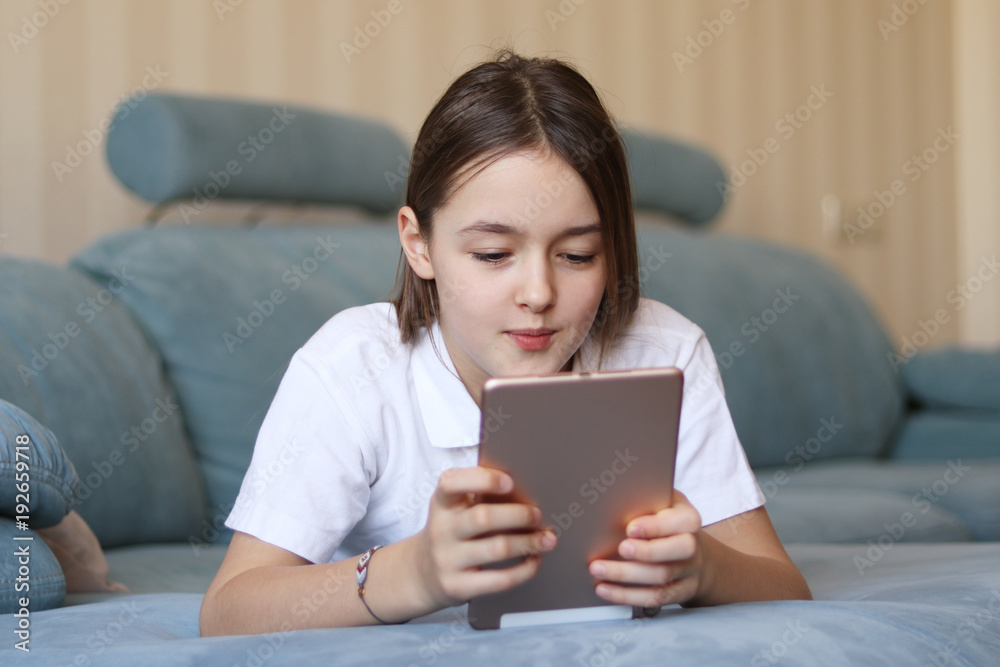 Beautiful preteen girl lying on sofa looking with interest at digital tablet pad in the living room at home. Child and gadget concept Beautiful preteen girl lying on sofa looking with interest at digital tablet pad in the living room at home. Child and gadget concept