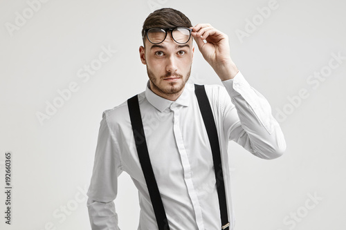 Human facial expressions and emotions. Portrait of emotional fashionable young bearded Caucasian male dressed in formal wear lifting his stylish eyeglasses, having confused questioning look