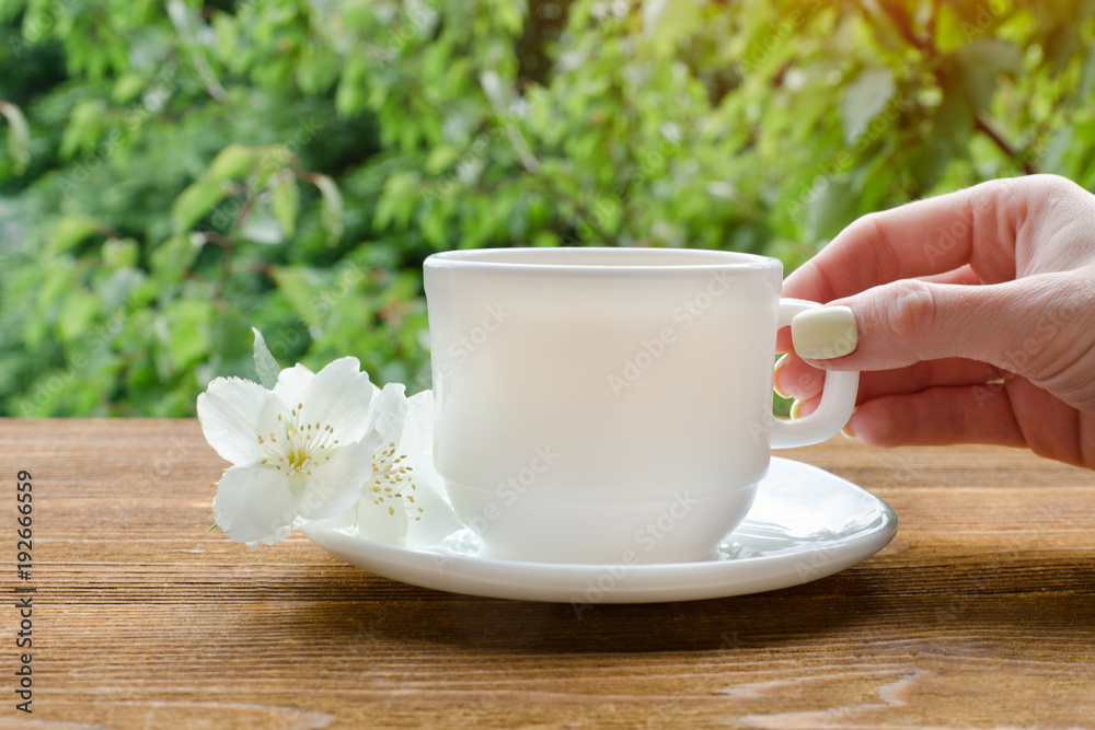 Female hand with white mug of tea and jasmine. Greenery on background.