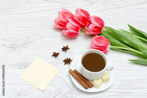 Pink tulips, mug of coffee and cinamon. Empty postcard. Llight wooden background.