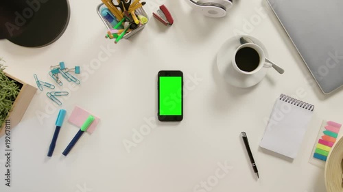 A finger touching a smartphone with a green screen. The phone is on the white table. View from the top. Close-up.