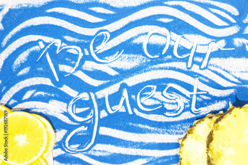 Be our guest inscription on a blue colored sand with waves, sliced orange, pineapple, top view, flat lay photo