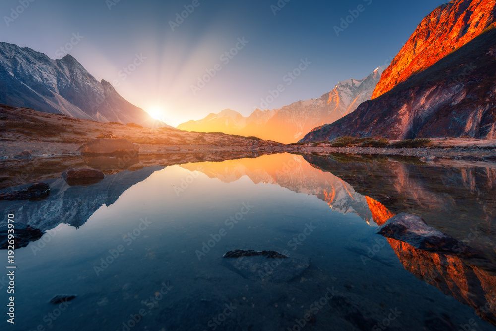 Fototapeta premium Piękny krajobraz z wysokimi górami z oświetlonymi szczytami, kamieniami w górskim jeziorze, odbiciem, błękitnym niebem i żółtym światłem słonecznym o wschodzie słońca. Nepal. Niesamowita scena z Himalajami. Himalaje
