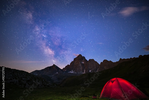 Camping under starry sky and milky way at high altitude on the Alps. Illuminated tent in the foreground and majestic mountain peak in the background. Adventure and exploration in summertime.