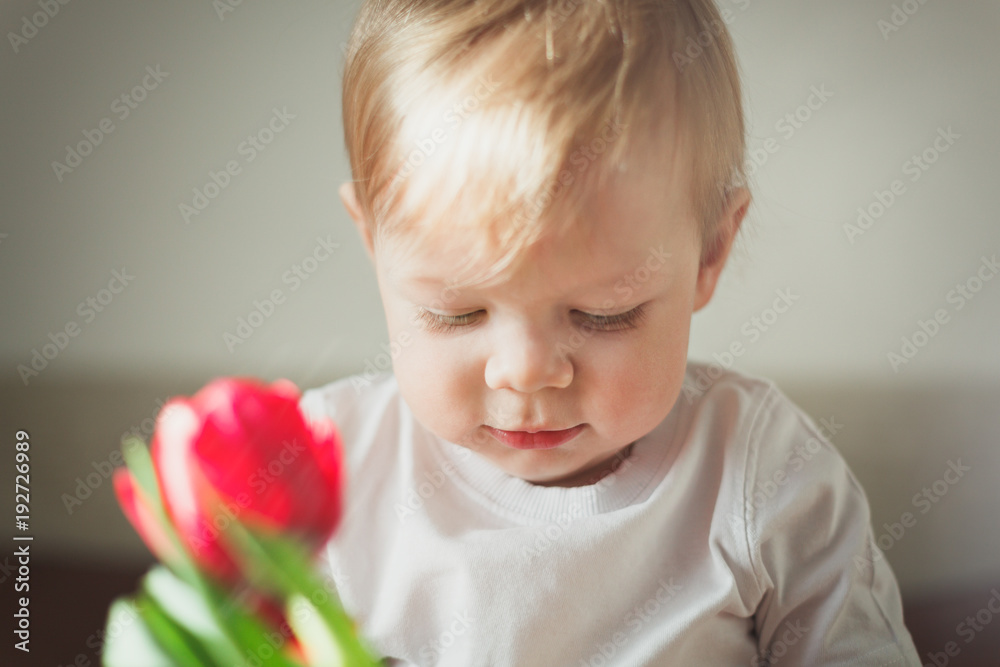 Portrait of a cute little smiling boy. Close up. In the foreground not in focus there is a red Tulip flower and a small glare. Warm colour scheme. Home and cozy atmosphere. Sunny day. Portrait of a cute little smiling boy. Close up. In the foreground not in focus there is a red Tulip flower and a small glare. Warm colour scheme. Home and cozy atmosphere. Sunny day.