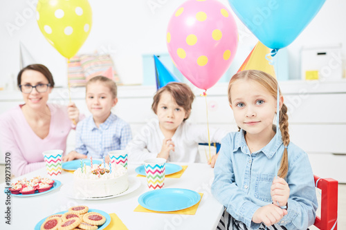 Cute child with pink balloon, her friends and teacher gathered by festive table at birthday party