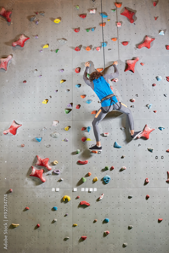 Sporty guy with climb equipment moving upwards while holding by rope and artificial ledges on climbing wall