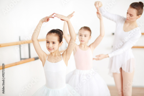 Little ballerina dancing with her arms over head while teacher helping her friend near by