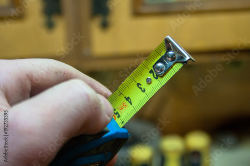 Marco focus is set on the yellow measuring tape that the man is holding in his hand. Workshop and tools in the background