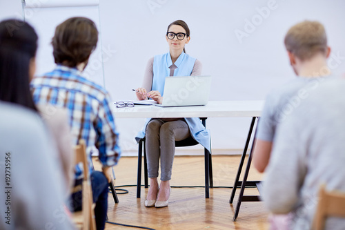 Young teacher sitting by desk in front of audience while making presentation at conference