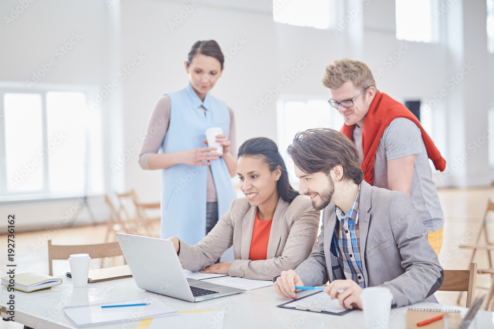 Several young associates in formalwear looking at laptop display and discussing online information at workplace